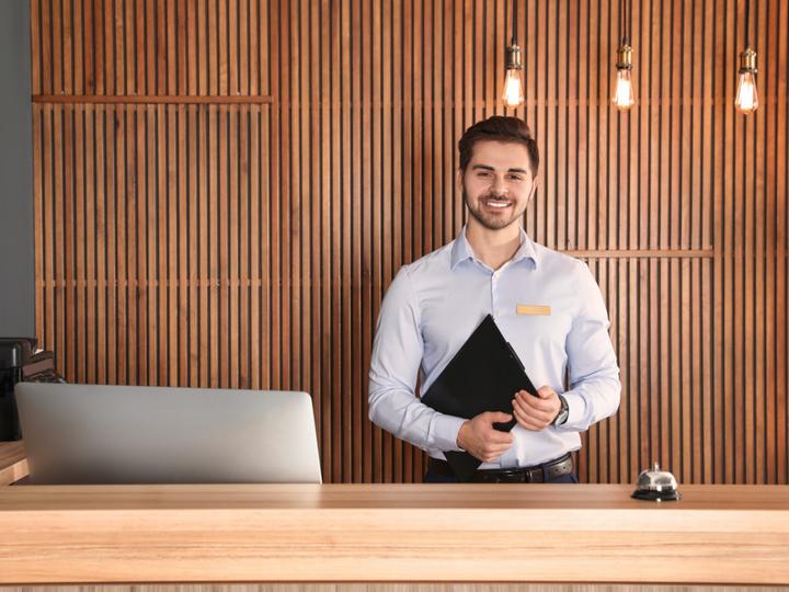 Frontoffice-Mitarbeiter steht mit einer Mappe in der Hand vor der Rezeptionstheke und lächelt.