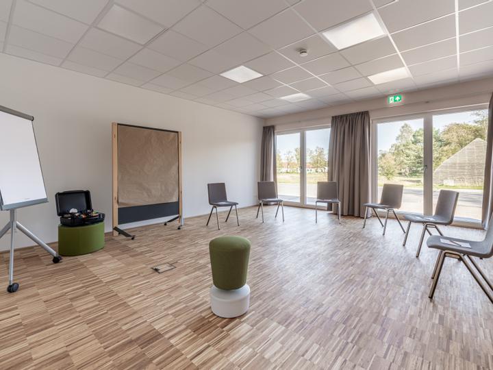 Tagungsraum mit Tisch und acht Stühlen. Raumhohe Fenster, Pinwand und Moderatorenkoffer