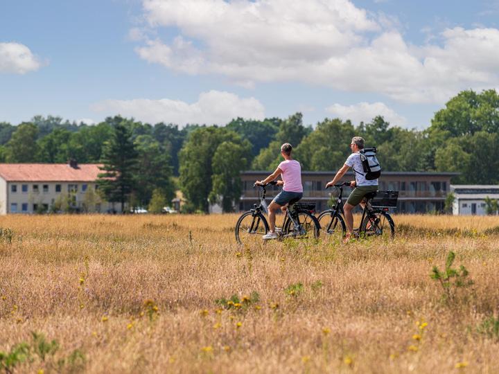 Zwei Radfahrer radeln durch eine offene Landschaft, im Hintergrund sind Hotelgebäude zu sehen.
