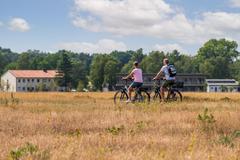 Zwei Radfahrer radeln durch eine offene Landschaft, im Hintergrund sind Hotelgebäude zu sehen.